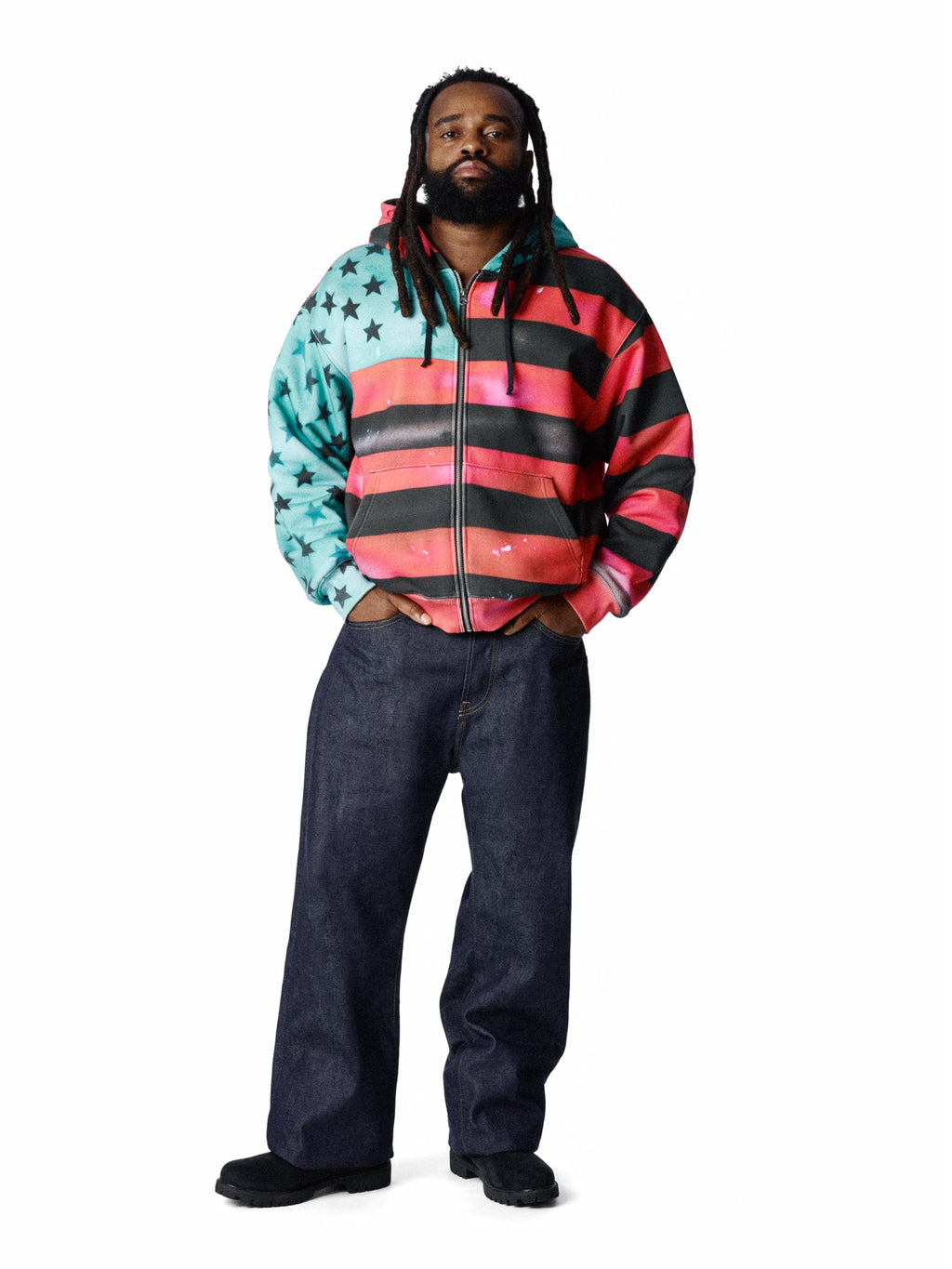 A man modeling a zip-up DENIM TEARS hoodie with a colorful American flag-inspired design, featuring a turquoise and black star-patterned left side, black and pink horizontal stripes on the right side, and a black zipper. He is wearing dark blue jeans and black shoes against a plain white background.