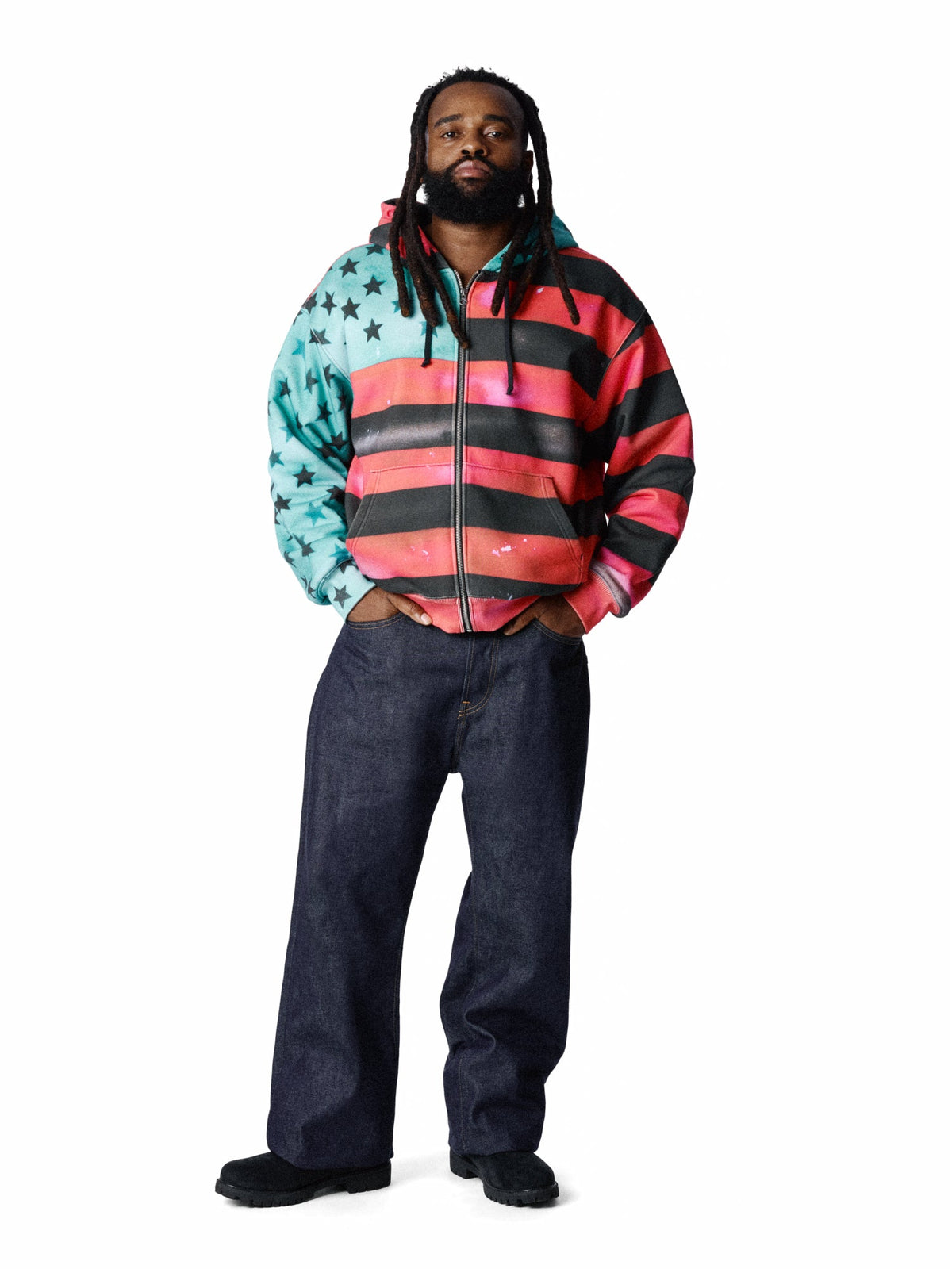 A man modeling a zip-up DENIM TEARS hoodie with a colorful American flag-inspired design, featuring a turquoise and black star-patterned left side, black and pink horizontal stripes on the right side, and a black zipper. He is wearing dark blue jeans and black shoes against a plain white background.