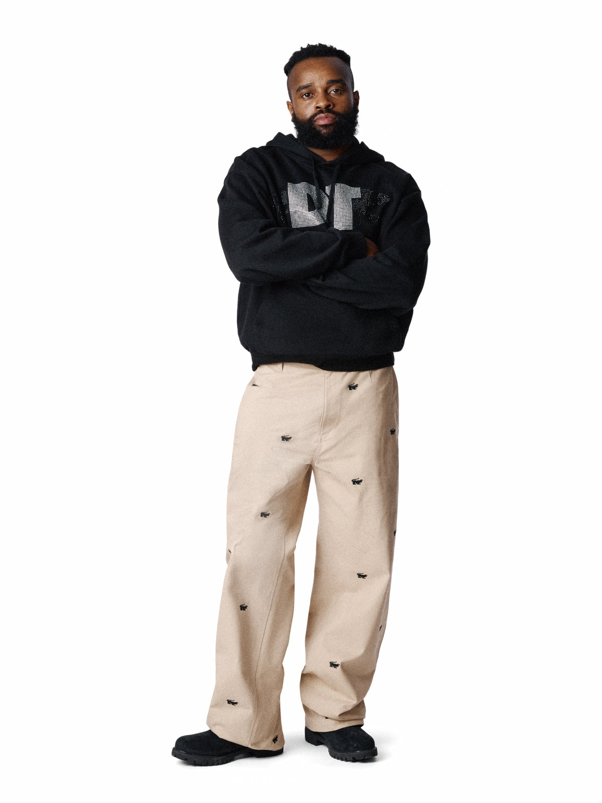 A man modeling a black Denim Tears hoodie with a gray graphic on the chest and beige pants with small black embroidered details, standing against a solid white background.