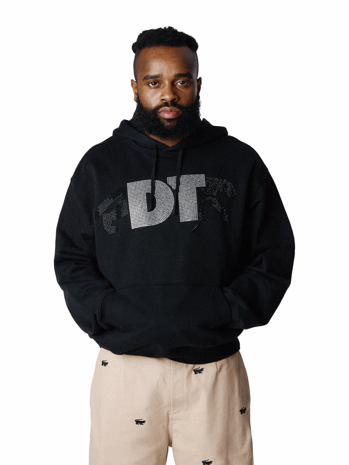 A man with a beard modeling a black pullover Denim Tears hoodie with a distressed silver 'DT' logo on the chest, paired with beige pants, against a solid white background.
