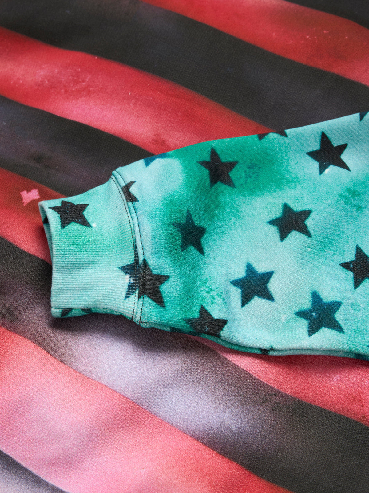 A close-up of a mint green DENIM TEARS Hoodie sleeve with a black star pattern, laid flat on a red and black striped fabric background.